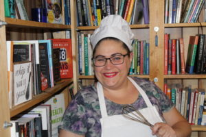 Photo of Jennifer Harshman, The Book Baker, wearing white hat and apron and holding a whisk